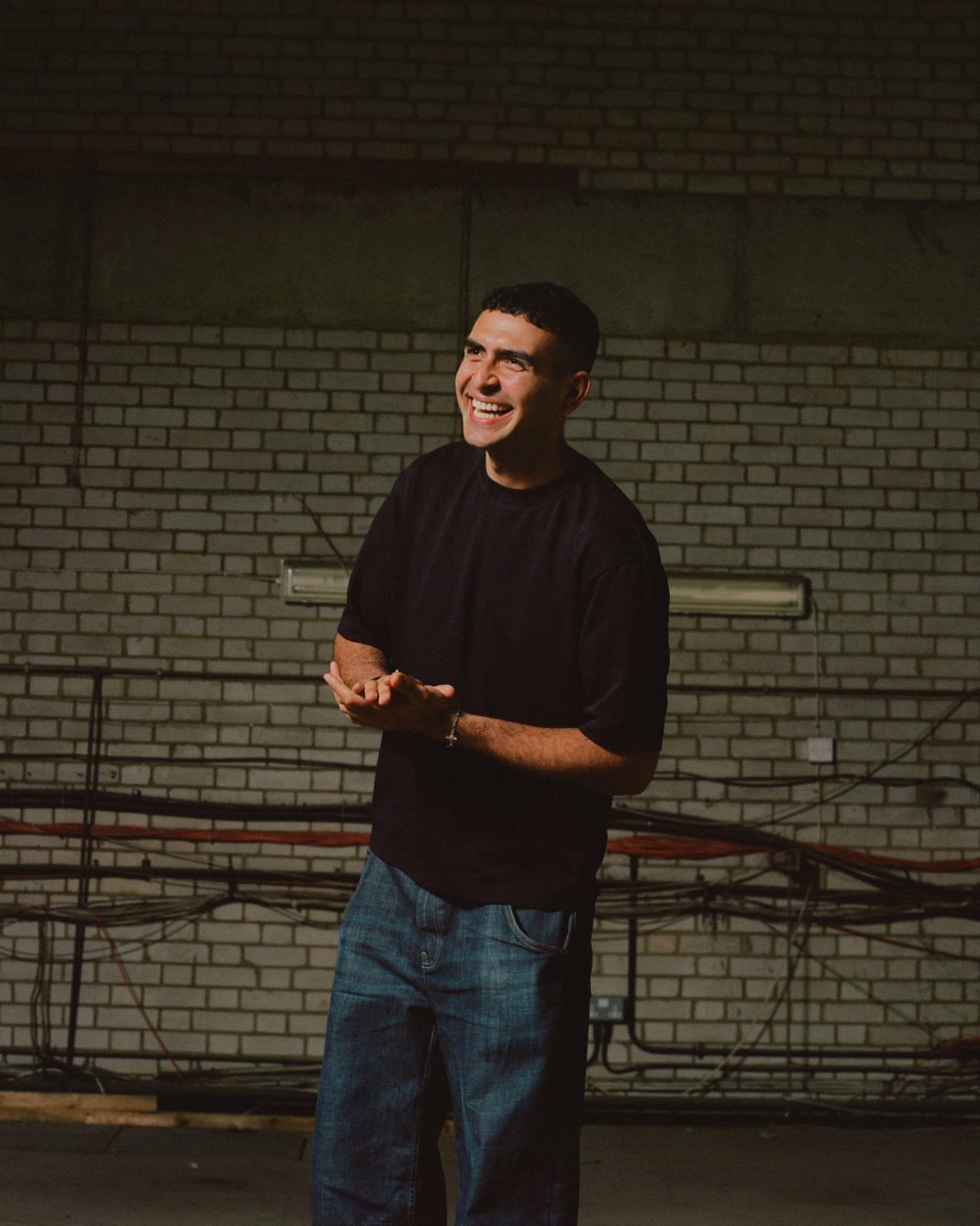 Sammy Virji laughing in a black t-shirt and jeans, standing in a dimly lit brick warehouse