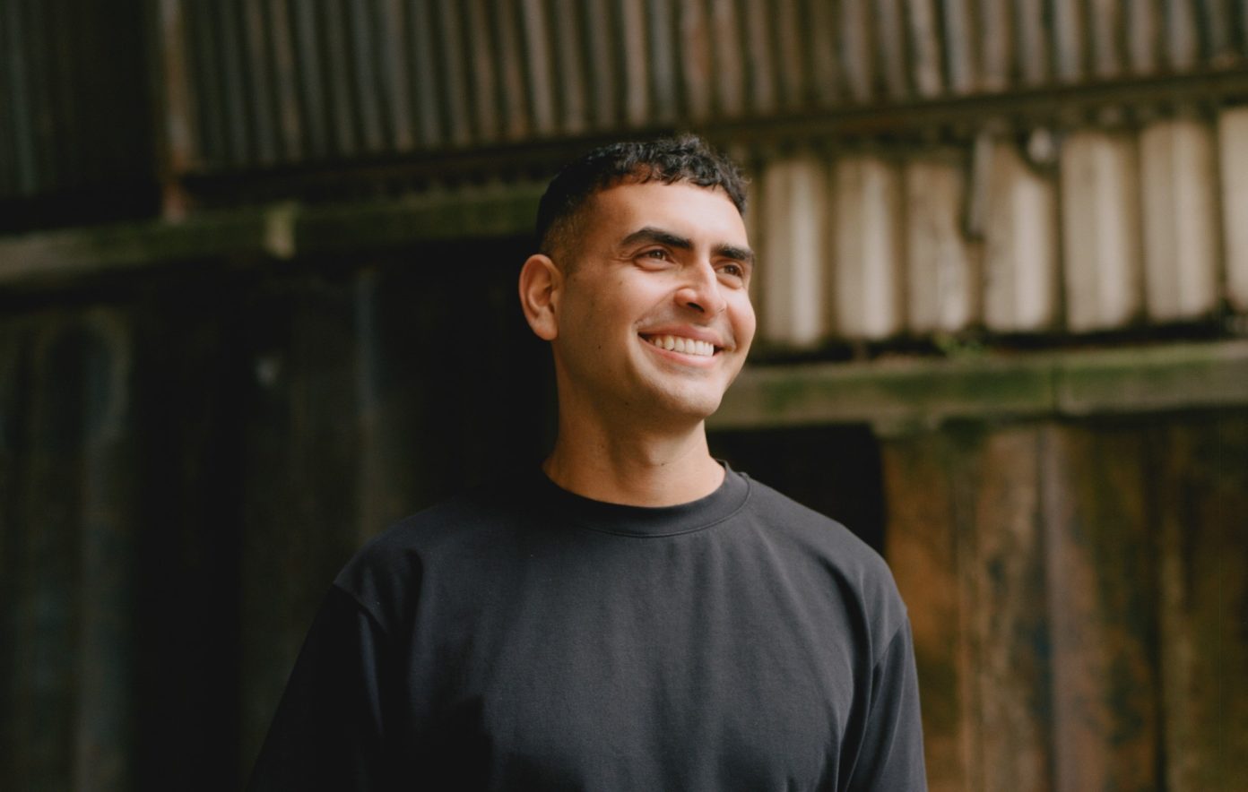Sammy Virji smiling, head and shoulders, in front of a weathered metal wall